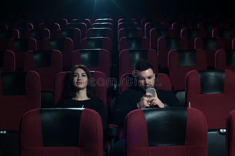 Young caucasian couple sitting in movie theater. Upset brunette girl watching film, while her boyfriend messaging using smartphone. Young caucasian couple sitting in movie theater. Upset brunette girl watching film, while her boyfriend messaging using smartphone.