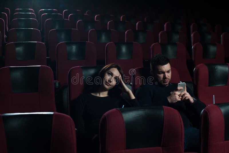 Young caucasian couple sitting in movie theater. Attractive brunette girl watching film, while her boyfriend messaging using smartphone. Cinema, entertainment and leisure concept. Young caucasian couple sitting in movie theater. Attractive brunette girl watching film, while her boyfriend messaging using smartphone. Cinema, entertainment and leisure concept.