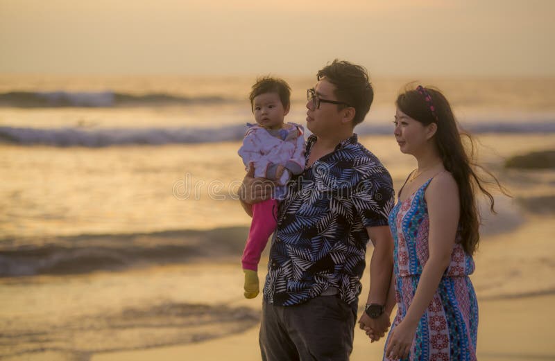 Jeunes couples coréens asiatiques heureux et beaux tenant la fille de bébé marchant sur la plage de coucher du soleil appréciant photographie stock libre de droits Jeunes couples coréens asiatiques heureux et beaux tenant la fille de bébé marchant sur la plage de coucher du soleil appréciant photographie stock libre de droits