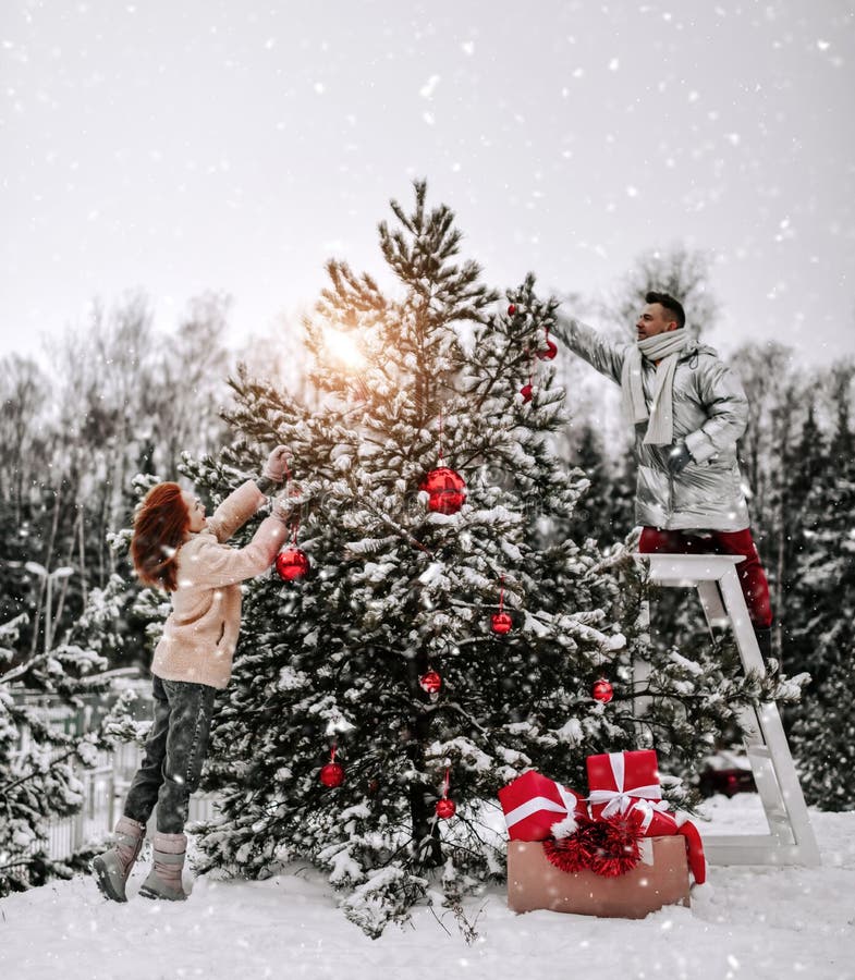 Jeunes couples heureux dans des vêtements d'hiver élégant décorent l'arbre de Noël avec des balles en plein air dans la forêt neig