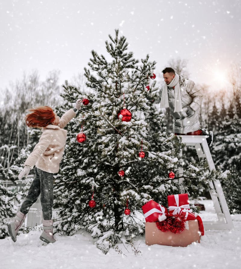 Jeunes couples heureux dans des vêtements d'hiver élégant décorent l'arbre de Noël avec des balles en plein air dans la forêt neig