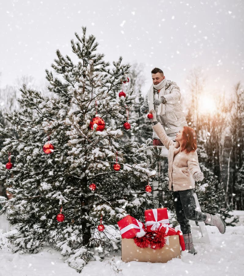 Jeunes couples heureux dans des vêtements d'hiver élégant décorent l'arbre de Noël avec les boules rouges dehors dans la forêt nei