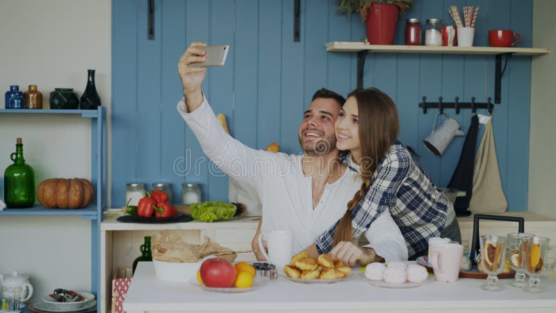 Jeunes couples heureux prenant le portrait de selfie tout en prenant le petit déjeuner dans la cuisine à la maison image libre de droits Jeunes couples heureux prenant le portrait de selfie tout en prenant le petit déjeuner dans la cuisine à la maison image libre de droits