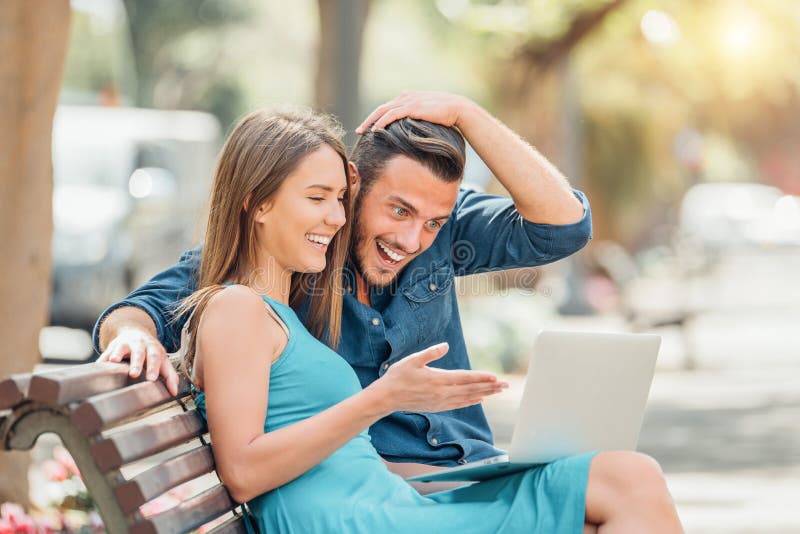Jeunes couples heureux utilisant l'ordinateur portable se reposant sur le banc dans la ville ext?rieure photographie stock Jeunes couples heureux utilisant l'ordinateur portable se reposant sur le banc dans la ville ext?rieure photographie stock