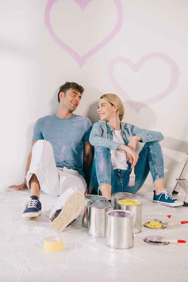 Happy young couple sitting on floor by white wall under drawn pink hearts. Happy young couple sitting on floor by white wall under drawn pink hearts