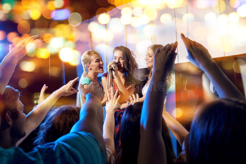Party, holidays, celebration, nightlife and people concept - happy young women singing karaoke in night club behind crowd of music fan. Party, holidays, celebration, nightlife and people concept - happy young women singing karaoke in night club behind crowd of music fan