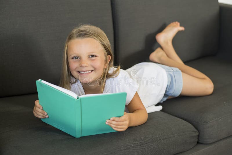 Jeunes petites années douces et heureuses de la fille 6 ou 7 se trouvant sur le divan à la maison de sofa de salon lisant un livr photos stock Jeunes petites années douces et heureuses de la fille 6 ou 7 se trouvant sur le divan à la maison de sofa de salon lisant un livr photos stock