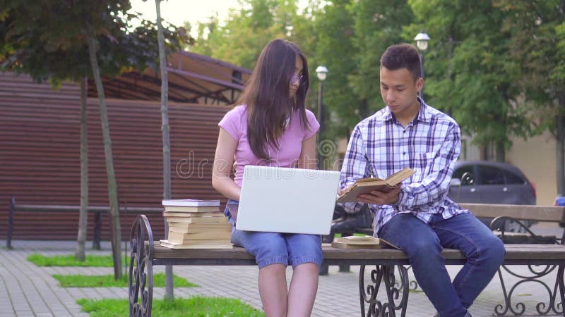 Jeunes Ã©tudiants asiatiques homme avec un livre et femme avec un ordinateur portable assis sur un banc lent