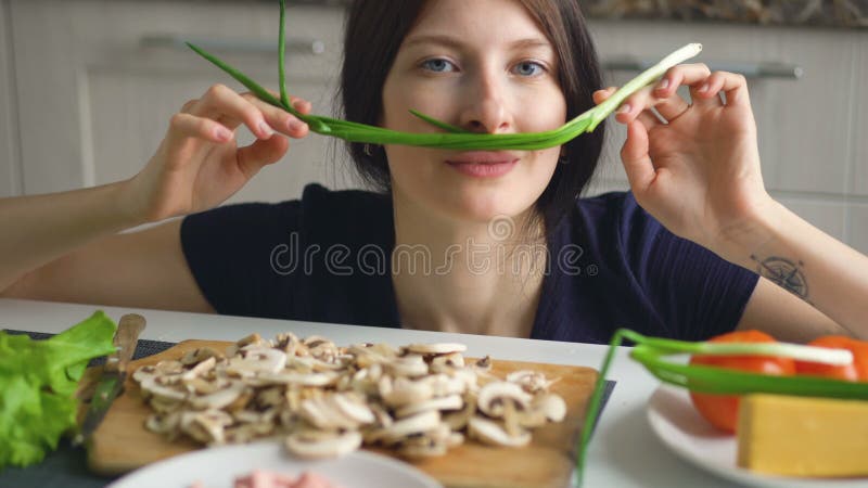 Jogo engraçado bonito do cozinheiro da mulher com a cebola verde na tabela na cozinha em casa