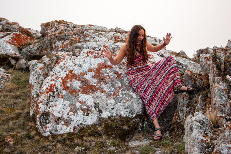 Cheerful pretty girl in a red striped dress with open mouth, arms wide open and legs lifted. Leans on a huge stone with red moss. European girl with long dark hair and a sly look. Cheerful pretty girl in a red striped dress with open mouth, arms wide open and legs lifted. Leans on a huge stone with red moss. European girl with long dark hair and a sly look