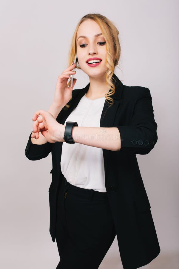Jolie jeune femme d'affaires joyeuse dans le costume de bureau parlant au téléphone, souriant et regardant la montre sur le fond photographie stock Jolie jeune femme d'affaires joyeuse dans le costume de bureau parlant au téléphone, souriant et regardant la montre sur le fond photographie stock