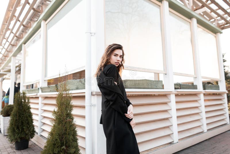 Jolie jeune femme moderne attirante dans un long manteau noir chic dans la pose près d'une maison en bois d'été dans la ville photographie stock libre de droits Jolie jeune femme moderne attirante dans un long manteau noir chic dans la pose près d'une maison en bois d'été dans la ville photographie stock libre de droits