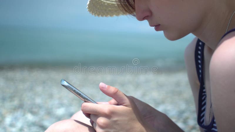 Jolie jeune femme à l'aide du smartphone par la plage de mer Fille dans le rétro chapeau et le maillot de bain avec les rayures b