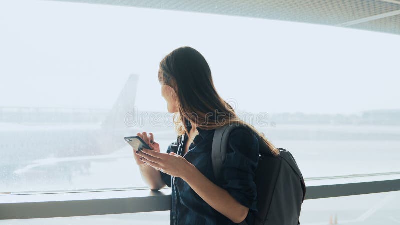 Jong meisje die smartphone gebruiken dichtbij luchthavenvenster De gelukkige Europese vrouw met rugzak gebruikt mobiele app in te royalty-vrije stock afbeeldingen Jong meisje die smartphone gebruiken dichtbij luchthavenvenster De gelukkige Europese vrouw met rugzak gebruikt mobiele app in te royalty-vrije stock afbeeldingen
