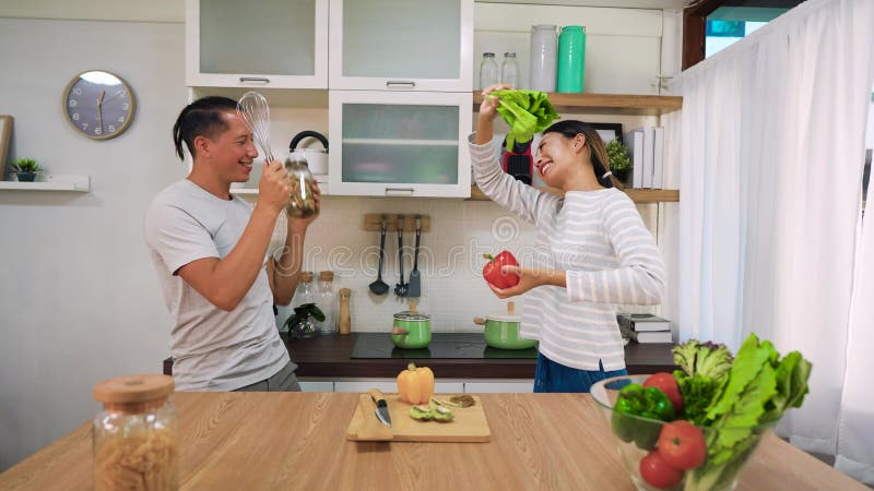 Jong mooi koppel danst in de keuken met raam. aziatische vrouw en kaukasiër die met groenten en fruit zingt