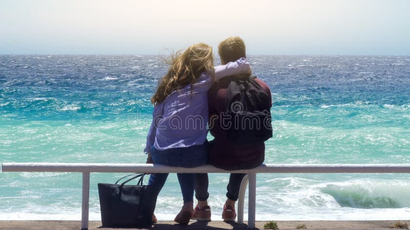 Jong paar die op de pijp van het wachtspoor op winderige zonnige dag koesteren, die stormachtige oceaan onder ogen zien stock fotografie Jong paar die op de pijp van het wachtspoor op winderige zonnige dag koesteren, die stormachtige oceaan onder ogen zien stock fotografie