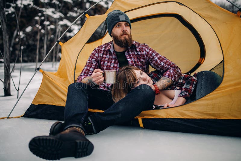 Young couple is in yellow tourist tent during winter hike. Guy drinks hot tea from metal mug, girl lies on guy`s knees. Young couple is in yellow tourist tent during winter hike. Guy drinks hot tea from metal mug, girl lies on guy`s knees.