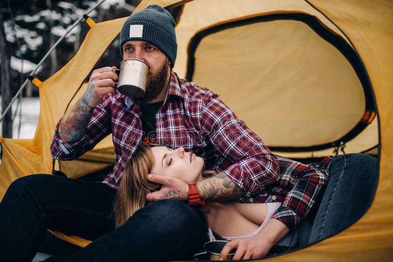 Young couple is in yellow tourist tent during winter hike. Guy drinks hot tea from metal mug, girl lies on guy`s knees. Closeup. Young couple is in yellow tourist tent during winter hike. Guy drinks hot tea from metal mug, girl lies on guy`s knees. Closeup.