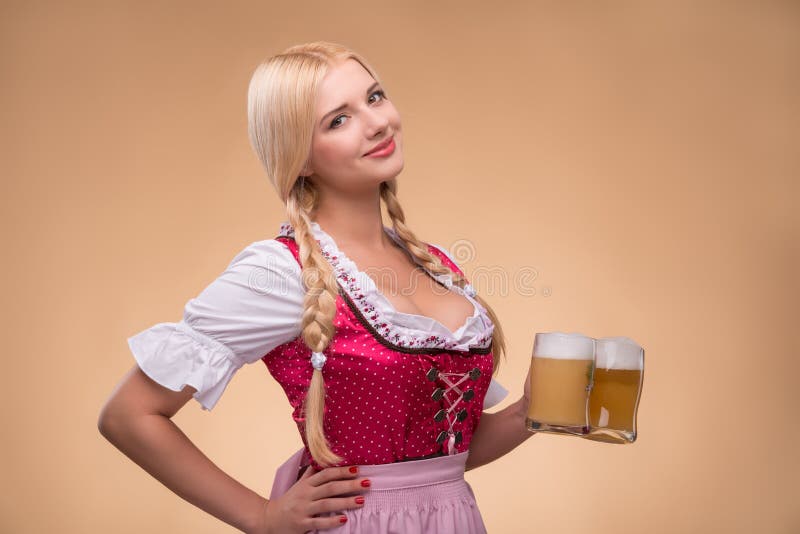 Half-length portrait of young smiling blonde wearing pink dirndl and white blouse standing aside holding in one hand beer mugs. Isolated on dark background. Half-length portrait of young smiling blonde wearing pink dirndl and white blouse standing aside holding in one hand beer mugs. Isolated on dark background