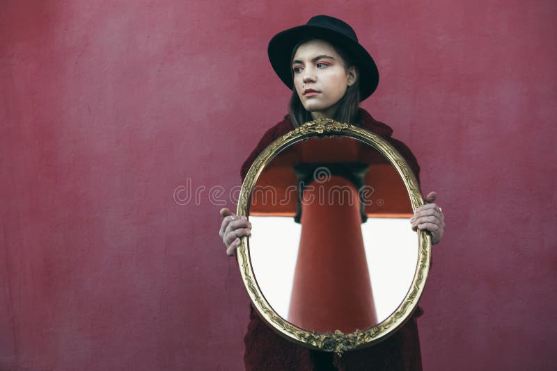 Jong tienermeisje in de spiegel van de hoedenholding voor rode muur de spiegel wijst op de kolom van het gebouw tegenover royalty-vrije stock foto Jong tienermeisje in de spiegel van de hoedenholding voor rode muur de spiegel wijst op de kolom van het gebouw tegenover royalty-vrije stock foto