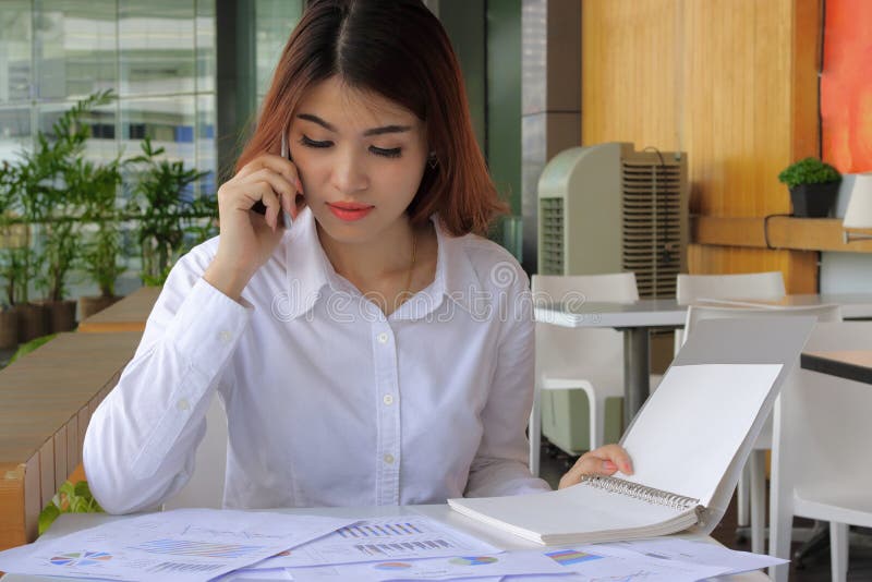 Jonge aantrekkelijke Aziatische bedrijfsvrouw die met mobiele telefoon, grafiek in bureau werken stock afbeelding Jonge aantrekkelijke Aziatische bedrijfsvrouw die met mobiele telefoon, grafiek in bureau werken stock afbeelding