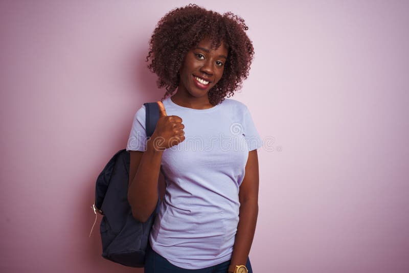 Young african afro tourist woman wearing backpack standing over isolated pink background doing happy thumbs up gesture with hand. Approving expression looking at the camera with showing success. Young african afro tourist woman wearing backpack standing over isolated pink background doing happy thumbs up gesture with hand. Approving expression looking at the camera with showing success
