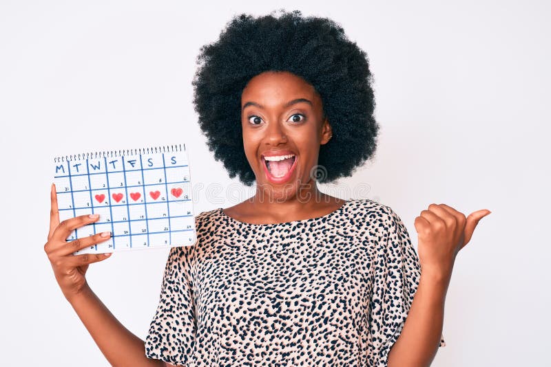 Young african american woman holding heart calendar pointing thumb up to the side smiling happy with open mouth. Young african american woman holding heart calendar pointing thumb up to the side smiling happy with open mouth