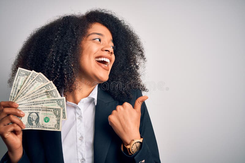 Young african american business woman with afro hair holding cash dollars banknotes pointing and showing with thumb up to the side with happy face smiling. Young african american business woman with afro hair holding cash dollars banknotes pointing and showing with thumb up to the side with happy face smiling