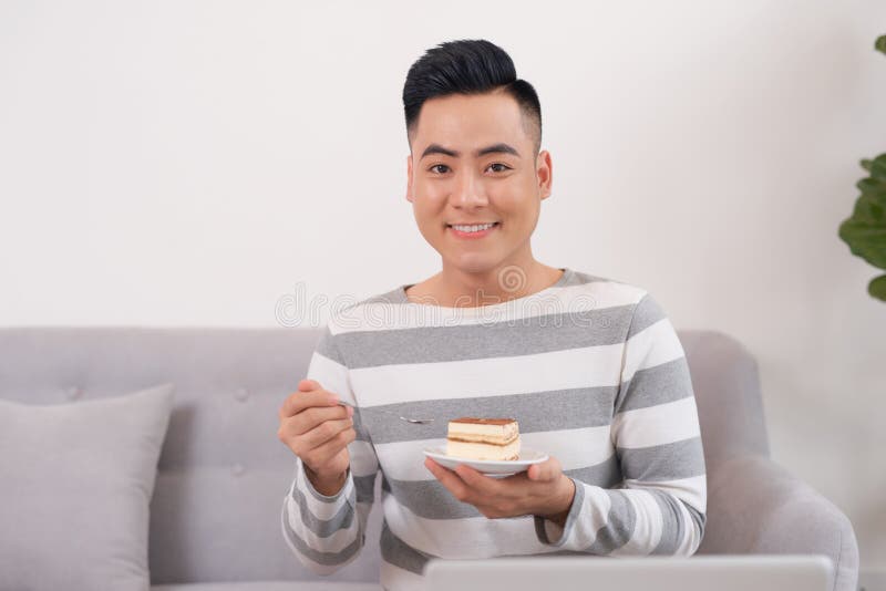 Jonge Aziatische mens die cake eten terwijl thuis het zitten op bank royalty-vrije stock afbeelding Jonge Aziatische mens die cake eten terwijl thuis het zitten op bank royalty-vrije stock afbeelding