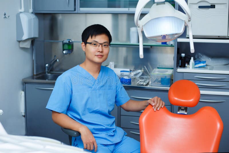 Jonge Aziatische mensentandarts met tandartsstoel bij de kliniek Dentis stock afbeelding Jonge Aziatische mensentandarts met tandartsstoel bij de kliniek Dentis stock afbeelding