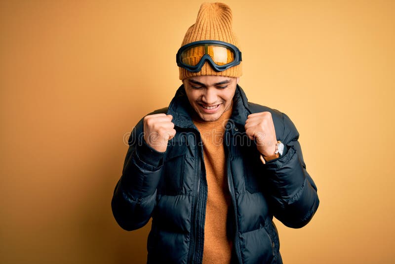 Young brazilian skier man wearing snow sportswear and ski goggles over yellow background excited for success with arms raised and eyes closed celebrating victory smiling. Winner concept. Young brazilian skier man wearing snow sportswear and ski goggles over yellow background excited for success with arms raised and eyes closed celebrating victory smiling. Winner concept