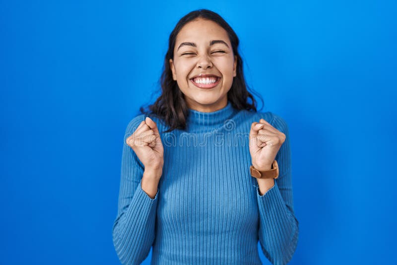 Young brazilian woman standing over blue isolated background excited for success with arms raised and eyes closed celebrating victory smiling. winner concept. Young brazilian woman standing over blue isolated background excited for success with arms raised and eyes closed celebrating victory smiling. winner concept