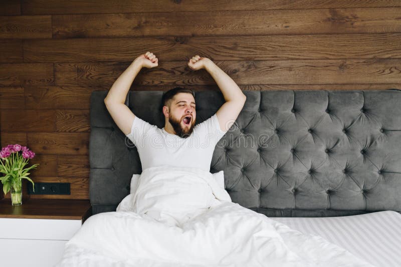 Jonge caucasiaanse bebaarde man die 's ochtends in zijn heldere moderne slaapkamer schreeuwt en rekt stock afbeelding Jonge caucasiaanse bebaarde man die 's ochtends in zijn heldere moderne slaapkamer schreeuwt en rekt stock afbeelding