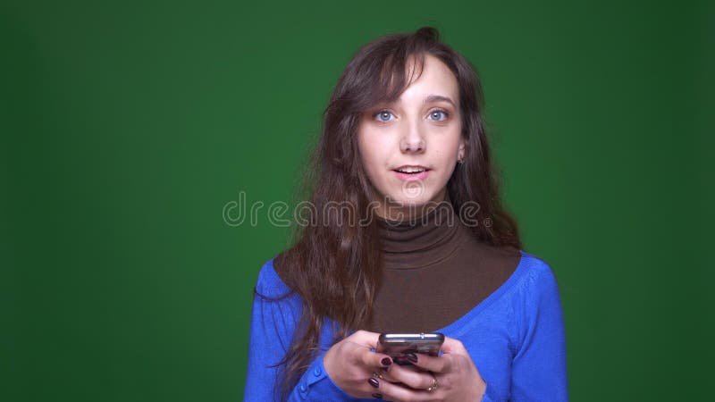 Jonge donkerbruine vrouwelijke student die smilingly met smartphone aan groene achtergrond werken