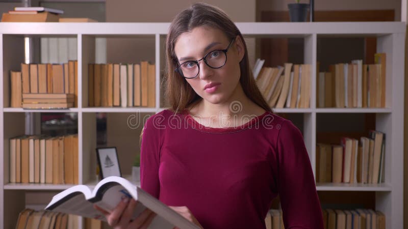 Jonge en mooie donkerbruine vrouwelijke student die in oogglazen boekhorloges lezen in camera in bibliotheek