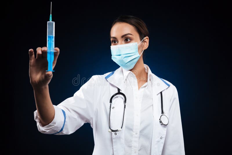 Jonge ernstige dame in medische masker holding en het bekijken spuit stock afbeeldingen Jonge ernstige dame in medische masker holding en het bekijken spuit stock afbeeldingen
