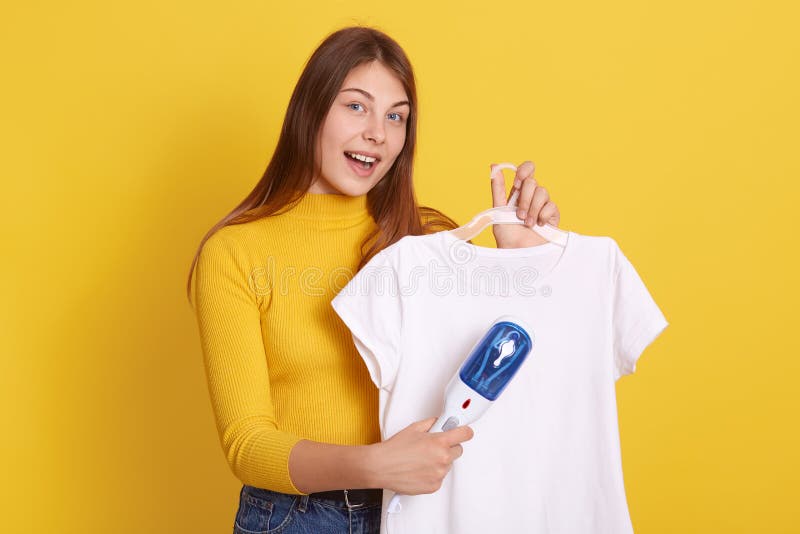 Jonge europese vrouw met een verbaasd strijken op shirt met een kleine stoomstrijkmachine kijkt naar een camera met een open mond