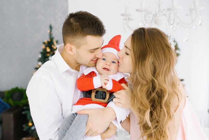 Jonge familie: het mamma en de papa kussen de wangen van de zoon van hun kind, die in een Santa Claus-kostuum gekleed is royalty-vrije stock foto's Jonge familie: het mamma en de papa kussen de wangen van de zoon van hun kind, die in een Santa Claus-kostuum gekleed is royalty-vrije stock foto's