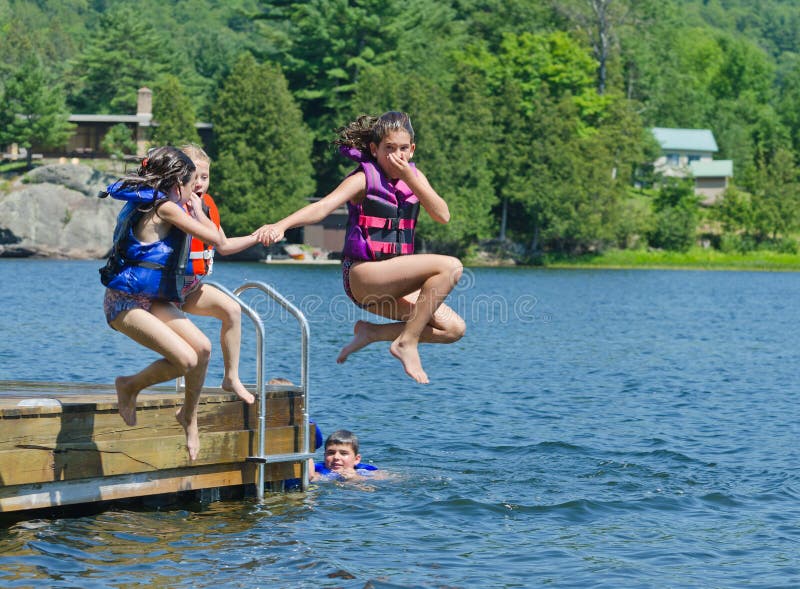 Kids having summer fun jumping off the dock into clear lake with life-jackets on. Kids having summer fun jumping off the dock into clear lake with life-jackets on