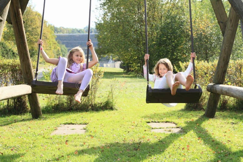 Barefoot kids - smiling girls in lila and white clothes sitting and swinging on swings. Barefoot kids - smiling girls in lila and white clothes sitting and swinging on swings