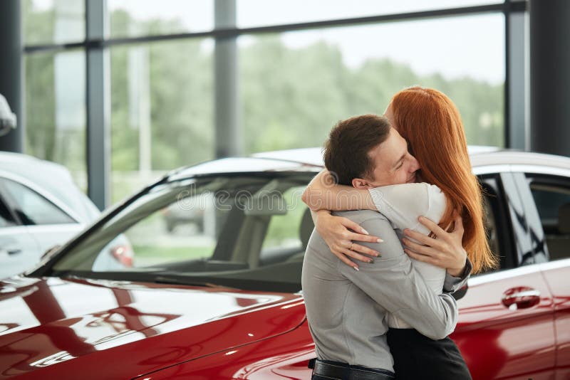 Jonge gelukkige die vrouw door een nieuwe auto bij autotoonzaal wordt verrast, gift van haar echtgenoot stock foto Jonge gelukkige die vrouw door een nieuwe auto bij autotoonzaal wordt verrast, gift van haar echtgenoot stock foto