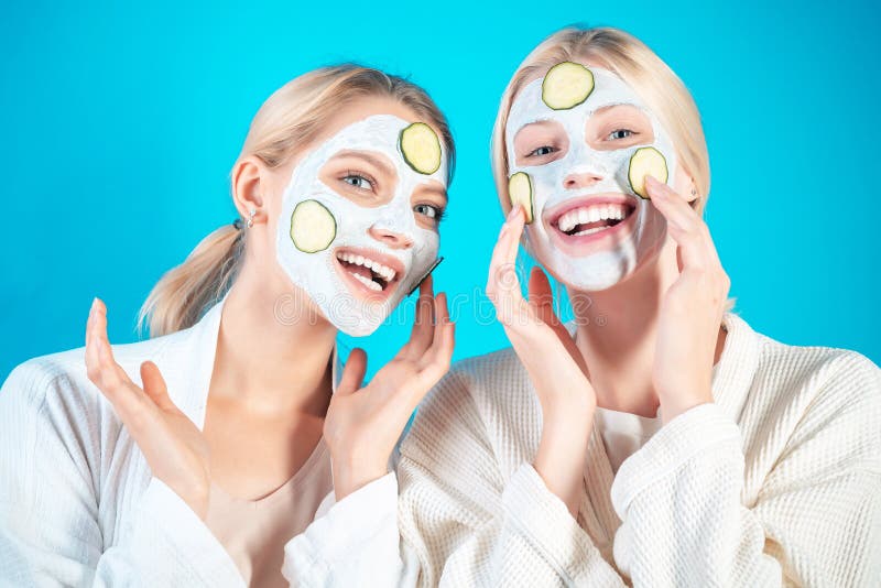 Young happy girls do facial mask with slices of cucumber at blue background. Skin care and treatment, spa, natural beauty and cosmetology concept. Young happy girls do facial mask with slices of cucumber at blue background. Skin care and treatment, spa, natural beauty and cosmetology concept