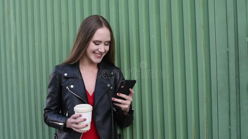 Jonge glimlachende blonde die kijkt naar haar smartphone en een glas koffie in haar hand