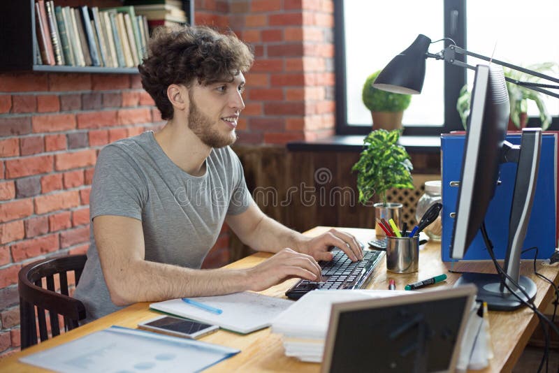 Jonge glimlachende mens die op het zijn werk aan bureaucomputer het typen bericht op toetsenbord in het bureau van de zolderstijl stock afbeeldingen Jonge glimlachende mens die op het zijn werk aan bureaucomputer het typen bericht op toetsenbord in het bureau van de zolderstijl stock afbeeldingen