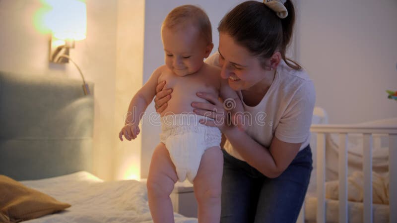 Jonge glimlachende moeder die zich 's nachts aan het kleden is en die haar babyzoon in bed laat zitten. begrip kinderhygiëne