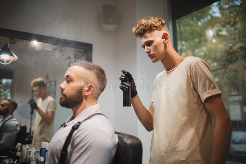 Jonge kapper met een haarlak die aan een vage achtergrond werken Modieuze cliënt in het concept van het de Kappersberoep van de k stock foto's Jonge kapper met een haarlak die aan een vage achtergrond werken Modieuze cliënt in het concept van het de Kappersberoep van de k stock foto's