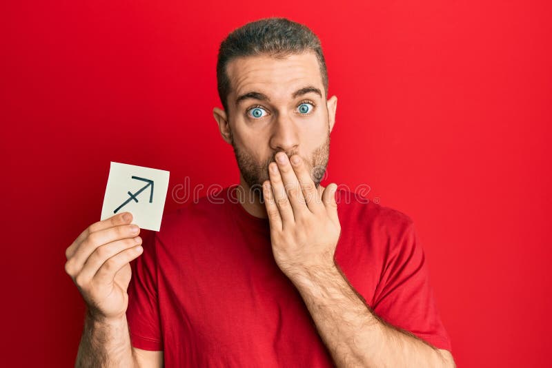 Young caucasian man holding paper with sagittarius zodiac sign covering mouth with hand, shocked and afraid for mistake. surprised expression. Young caucasian man holding paper with sagittarius zodiac sign covering mouth with hand, shocked and afraid for mistake. surprised expression