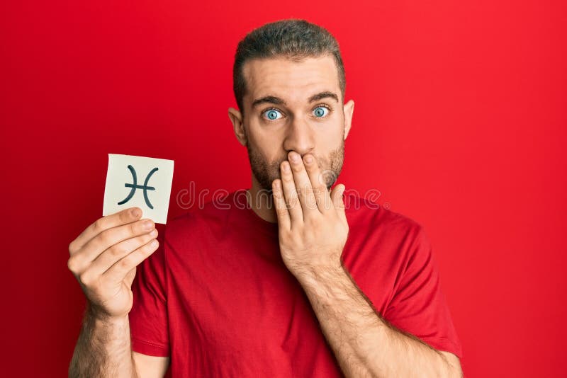 Young caucasian man holding paper with pisces zodiac sign covering mouth with hand, shocked and afraid for mistake. surprised expression. Young caucasian man holding paper with pisces zodiac sign covering mouth with hand, shocked and afraid for mistake. surprised expression