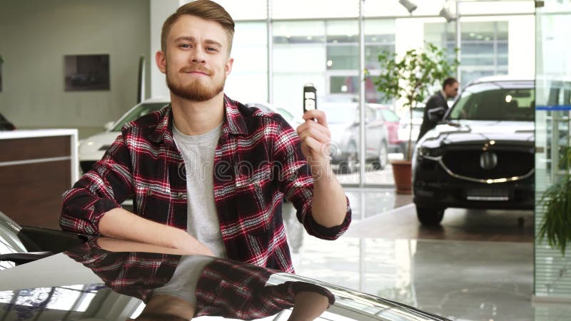 Jonge knappe mens die autosleutels tonen aan zijn nieuwe auto bij de het handel drijvensalon