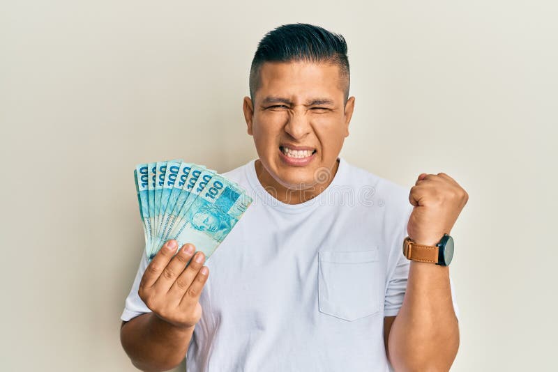 Young latin man holding 100 brazilian real banknotes screaming proud, celebrating victory and success very excited with raised arm. Young latin man holding 100 brazilian real banknotes screaming proud, celebrating victory and success very excited with raised arm